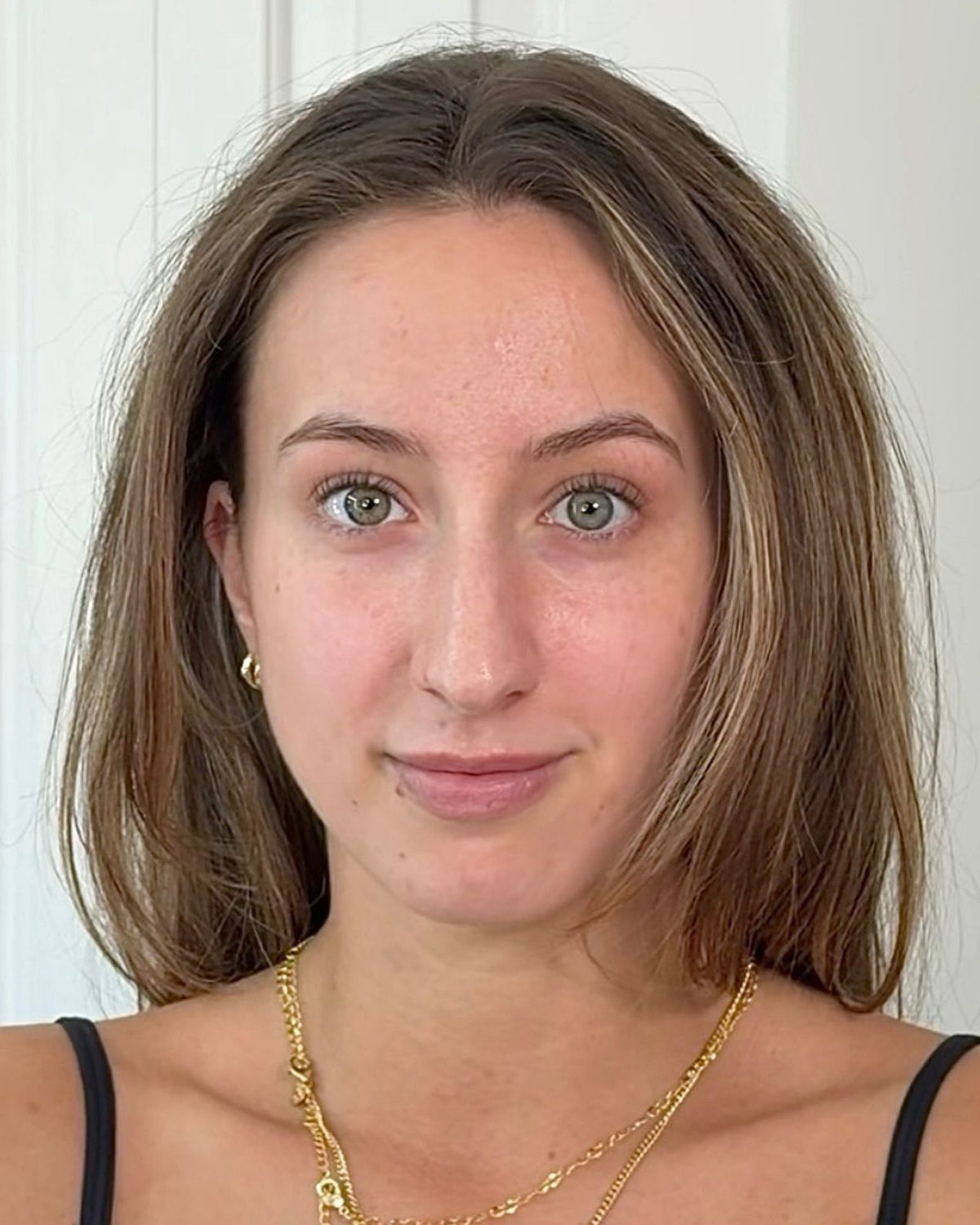 A close-up portrait of a woman with light brown hair, wearing a gold chain necklace and a small gold stud earring, against a white paneled background.
