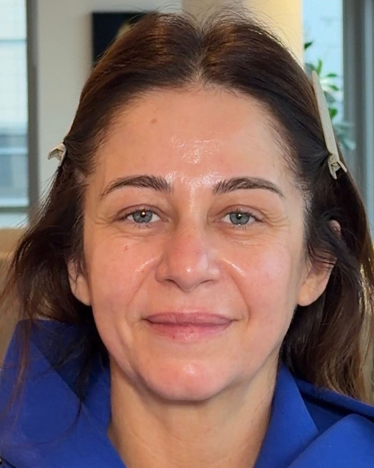 A close-up portrait of a woman with brown hair and blue eyes, wearing a blue top, photographed indoors with a softly blurred background.