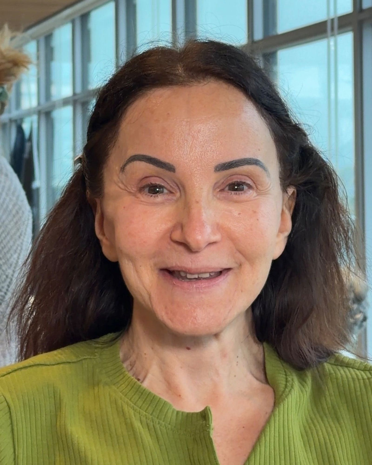 A close-up portrait of a smiling woman with dark brown hair wearing a textured olive green top, set against a bright window background.